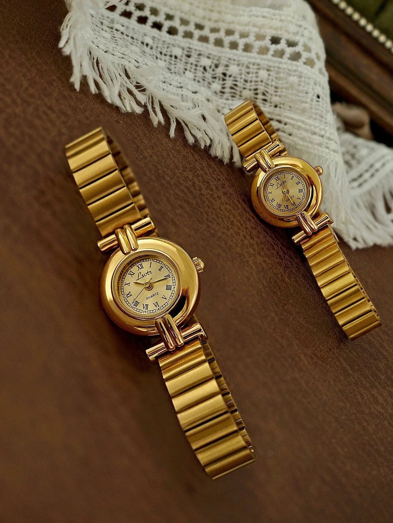 Two gold watches on a brown surface with a white lace fabric in the background.