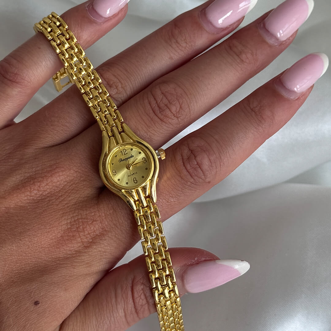 Gold watch on a hand with pink nail polish against a white background
