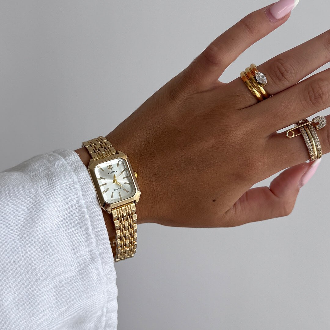 Hand wearing a gold watch and multiple rings on a plain background
