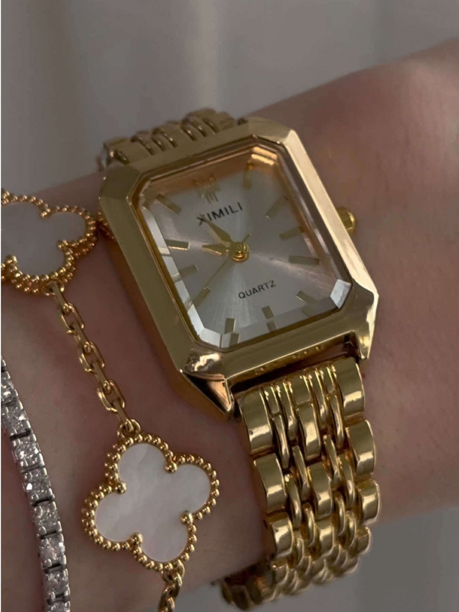 Gold wristwatch with a white face on a wrist, wearing a gold chain bracelet.