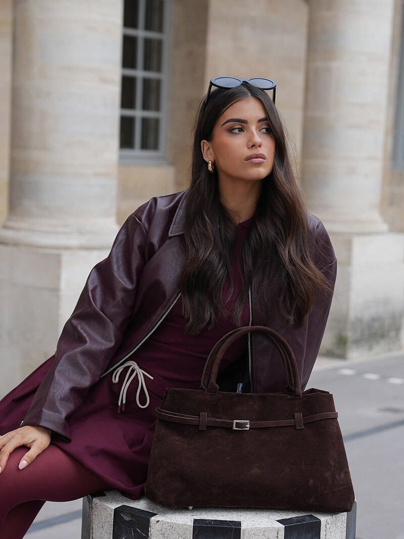 Woman in a burgundy coat with a brown handbag sitting on a stone bench.